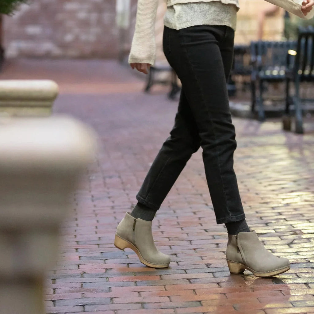 Person wearing black pants and beige Dansko Lizanne ankle boots on a brick pavement.