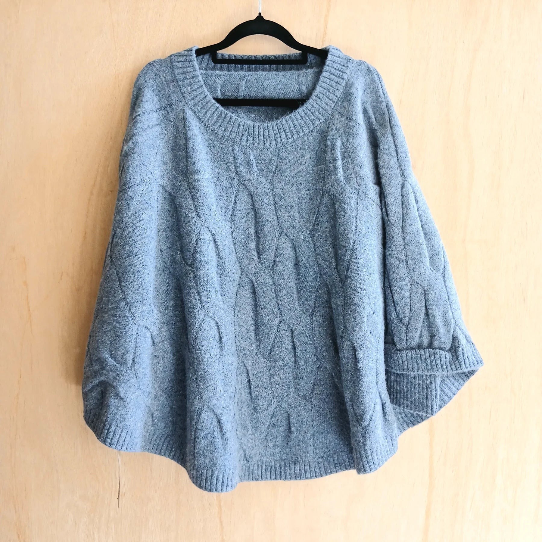The Happy Shoe Shop Cinnamon Creations blue chunky knit oversized poncho with cable texture on a black hanger.