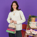The Happy Shoe Shop Vendula London book stack handbag held by a woman in a cream sweater against a purple background.