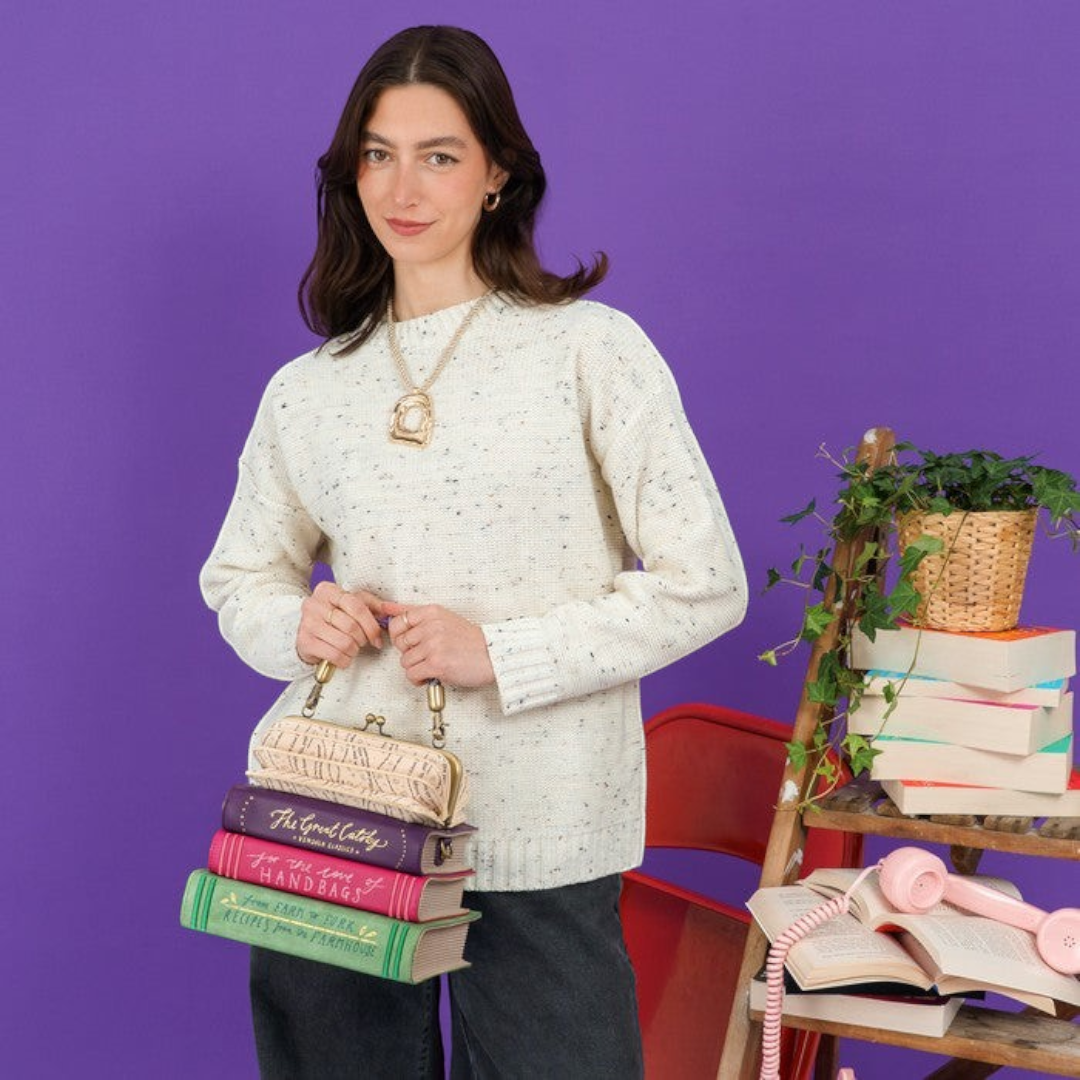 The Happy Shoe Shop Vendula London book stack handbag held by a woman in a cream sweater against a purple background.