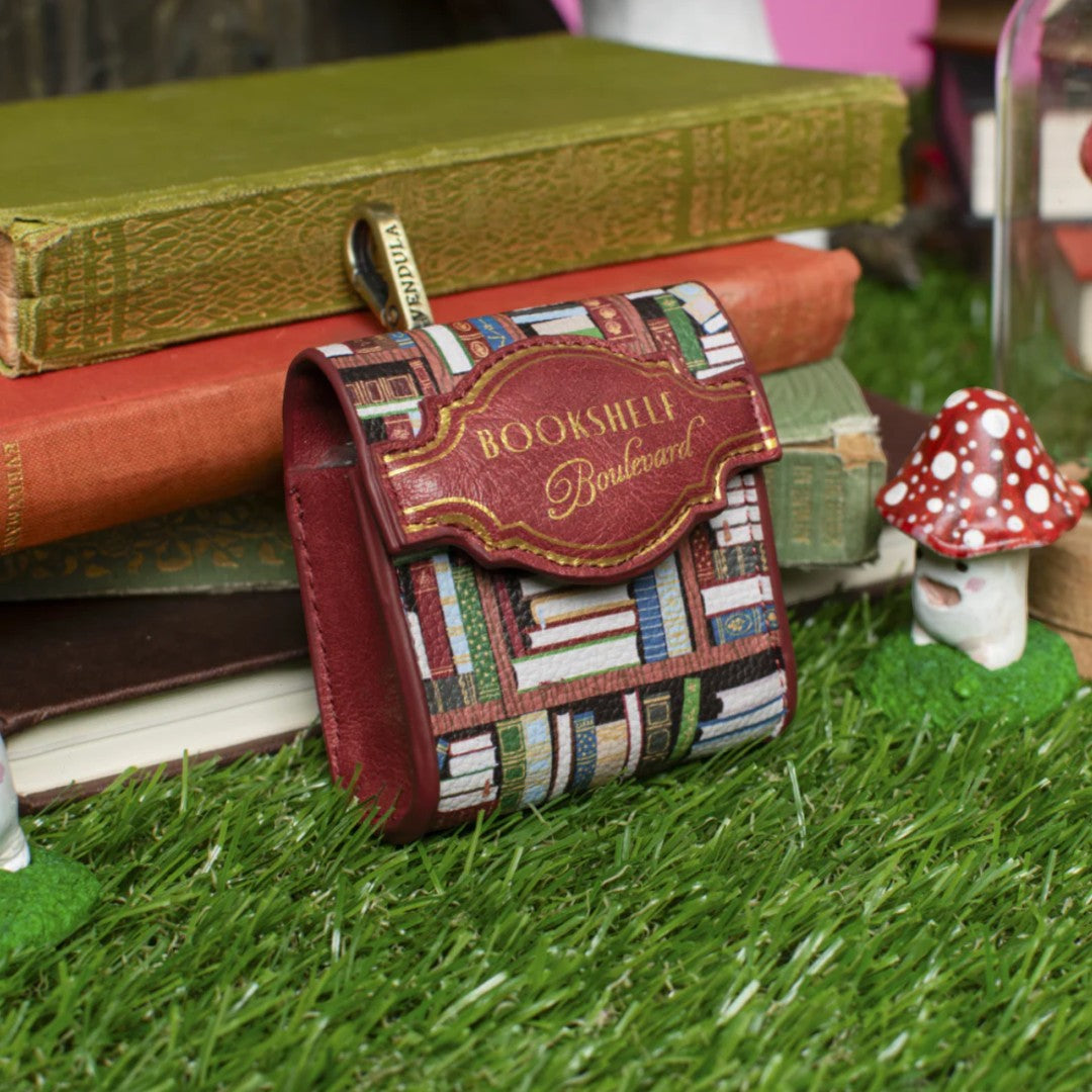 The Happy Shoe Shop Bookshelf Boulevard Pods Case, a small vegan leather pouch with a whimsical bookshelf print and a burgundy flap featuring "Bookshelf Boulevard" in gold lettering.