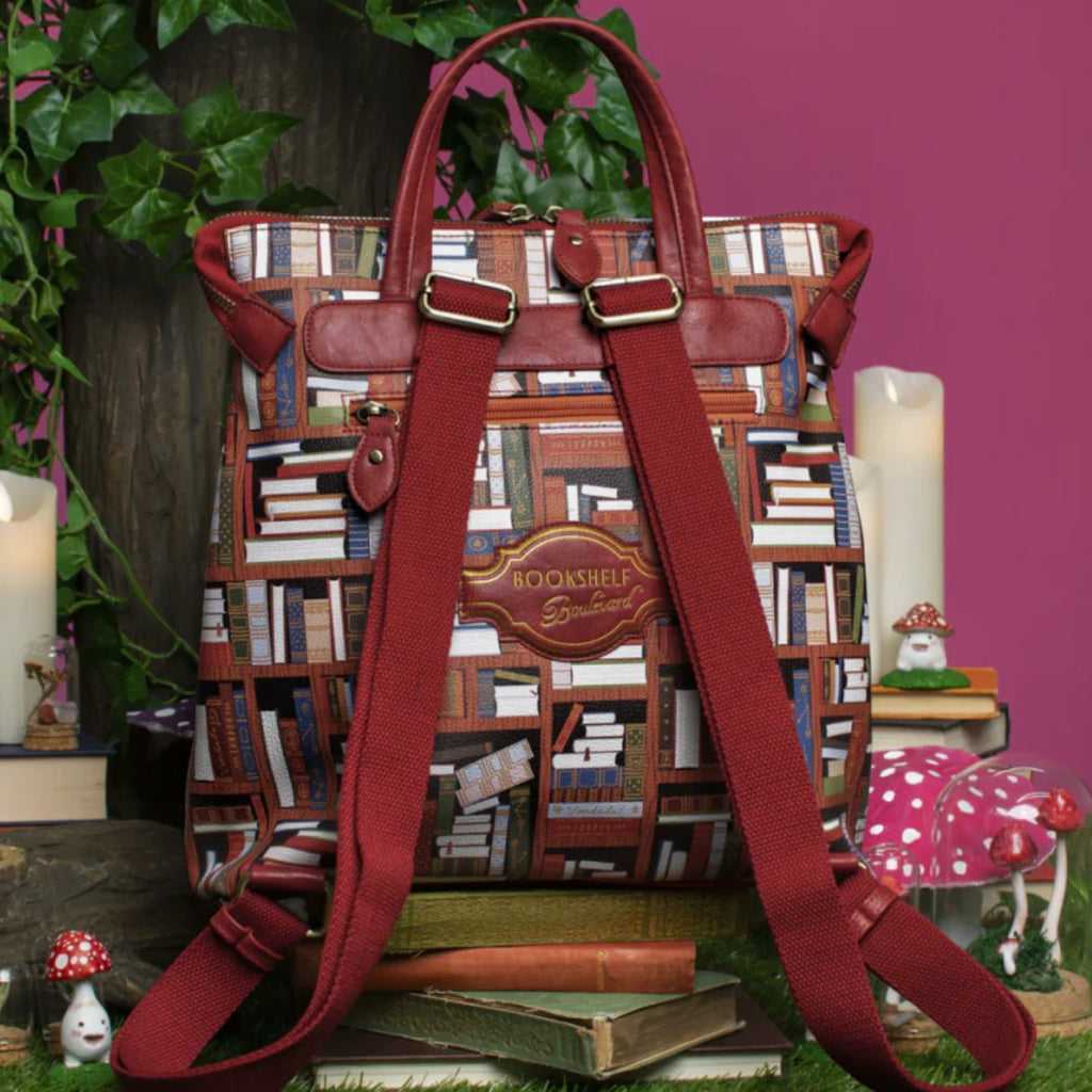 The Happy Shoe Shop Bookshelf Boulevard Tessa Backpack features a bookshelf print, vegan leather trim, and adjustable straps. It has a top handle and multiple pockets.