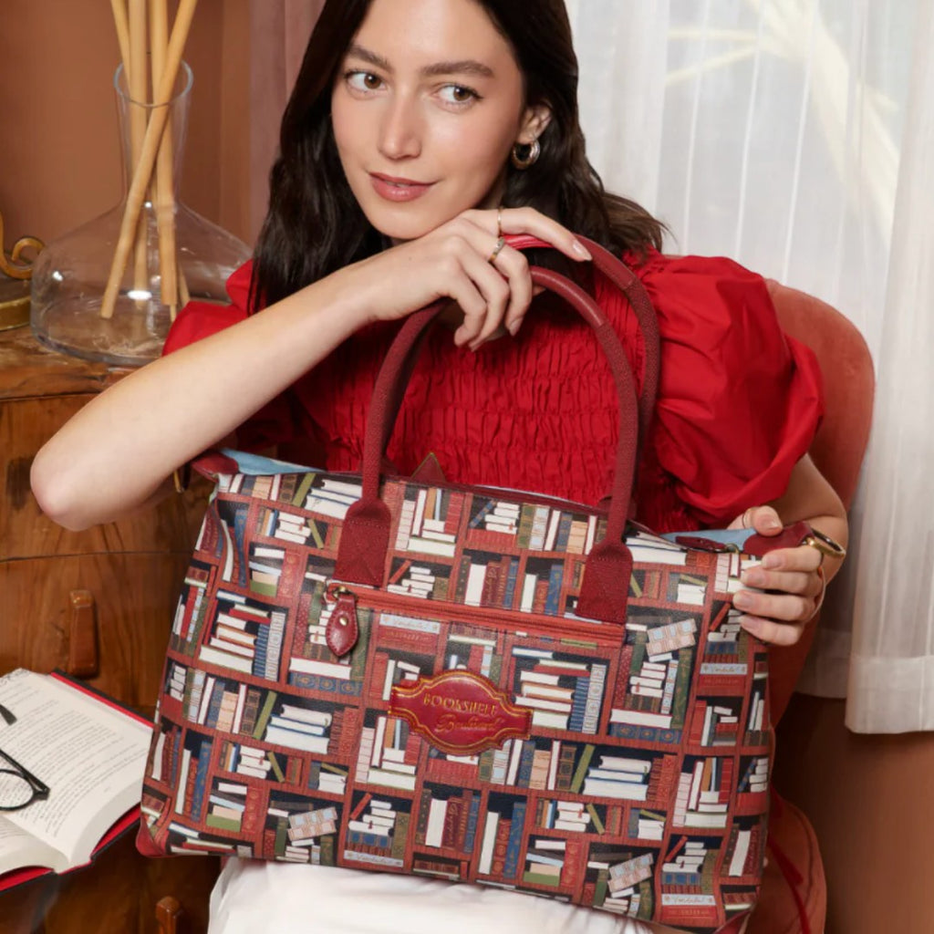 The Happy Shoe Shop Bookshelf Boulevard Bonnie Weekender Tote features a playful book-themed print on vegan leather with dual handles and a detachable strap.
