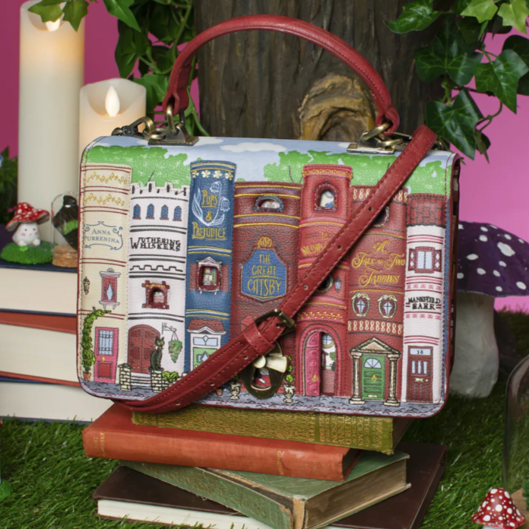The Happy Shoe Shop Bookshelf Boulevard Cassie Bag features a cityscape with book spines and dollhouse interiors, made of vegan leather with a red strap.