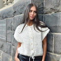 The Happy Shoe Shop cream sleeveless knit crop top with button front and adjustable drawstring hem on a model.