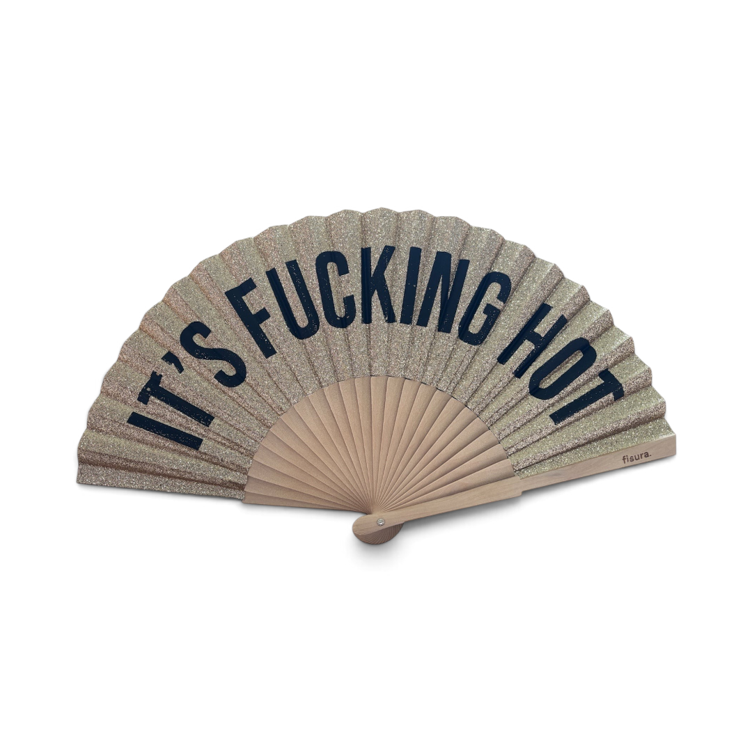 The Happy Shoe Shop Fisura hand fan in glittery gold with bold black text reading IT'S FUCKING HOT on bamboo ribs.