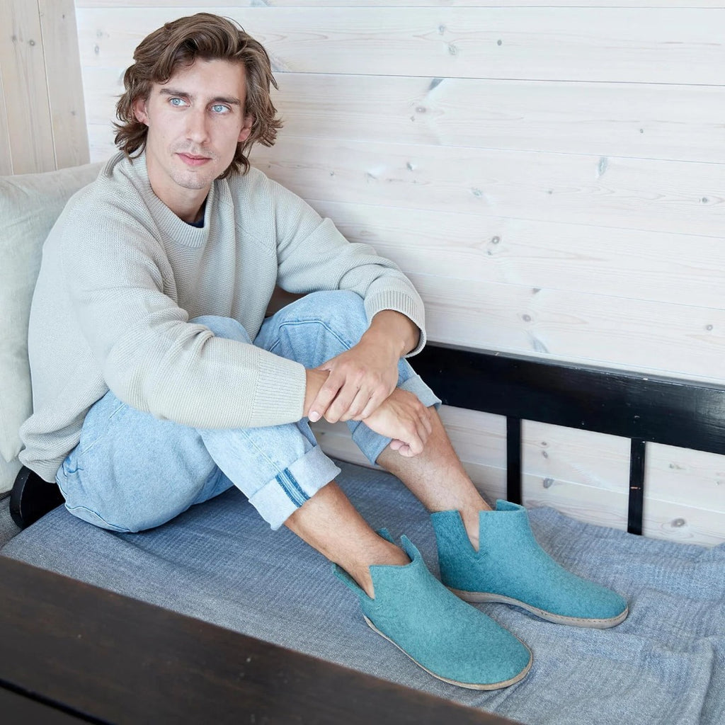 The Happy Shoe Shop Glerups The Boot, teal wool felt ankle boots with natural rubber sole, worn by a man sitting indoors.
