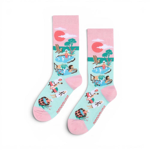 The Happy Shoe Shop Women's Hot Springs bamboo socks in light blue with pink cuffs, featuring a spa scene with people relaxing in pools and lounging chairs.