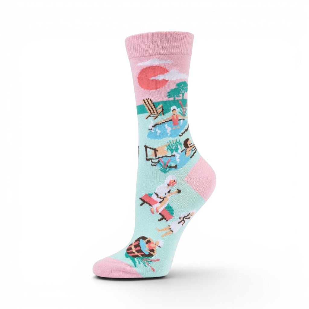 The Happy Shoe Shop Women's Hot Springs bamboo socks in light blue with pink trim, featuring a spa scene with people relaxing by a pool.