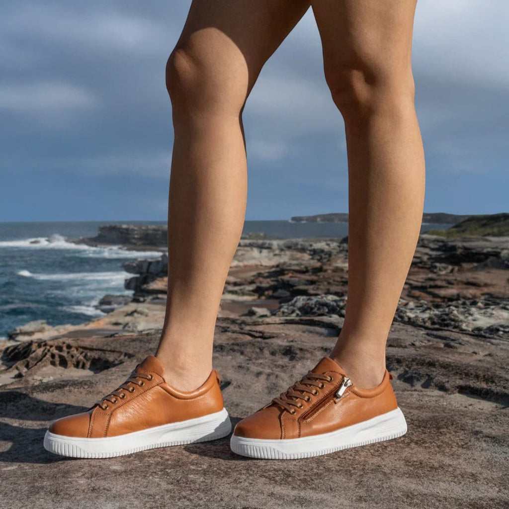 The Happy Shoe Shop Nelly tan leather sneakers with white platform soles and zip detail worn on a rocky coastal path.