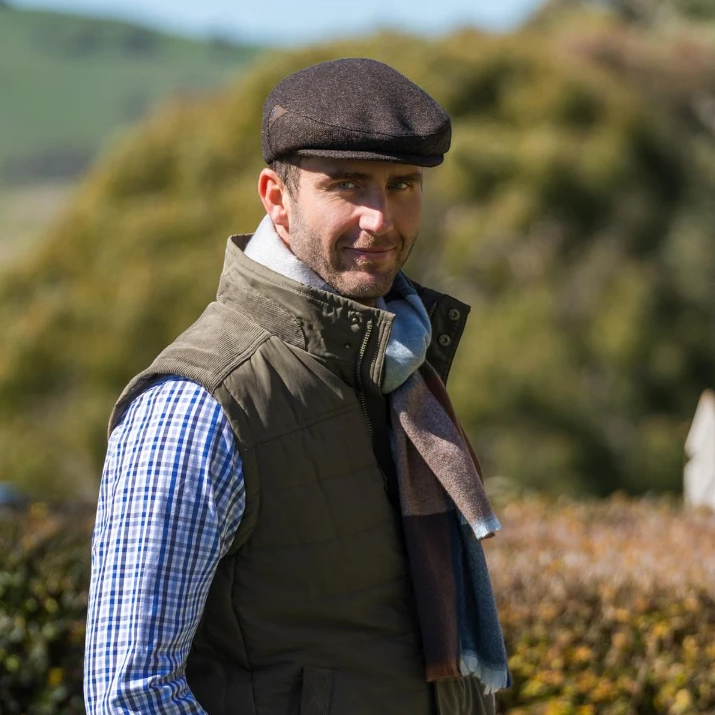 Thomas Cook Samuel Driver Cap in dark brown wool with a subtle embossed logo trim. Worn by a man outdoors.