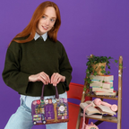 The Happy Shoe Shop Vendula London bowler bag featuring a bookshop print, held by a woman against a purple background.