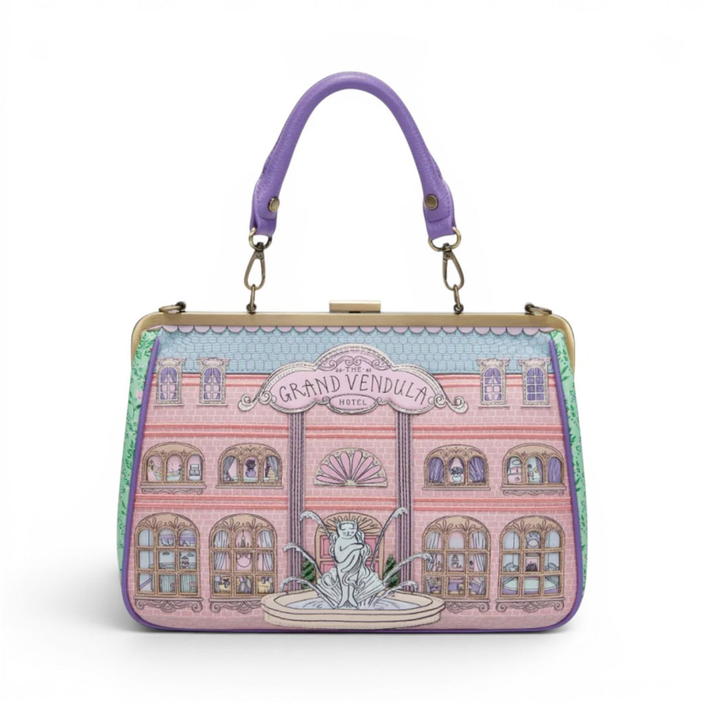 The Happy Shoe Shop Grand Vendula Hotel Willow Clip Frame Bag, pink vegan leather with hotel facade, purple handle, antique gold hardware.