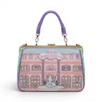 The Happy Shoe Shop Grand Vendula Hotel Willow Clip Frame Bag, pink vegan leather with hotel facade, purple handle, antique gold hardware.