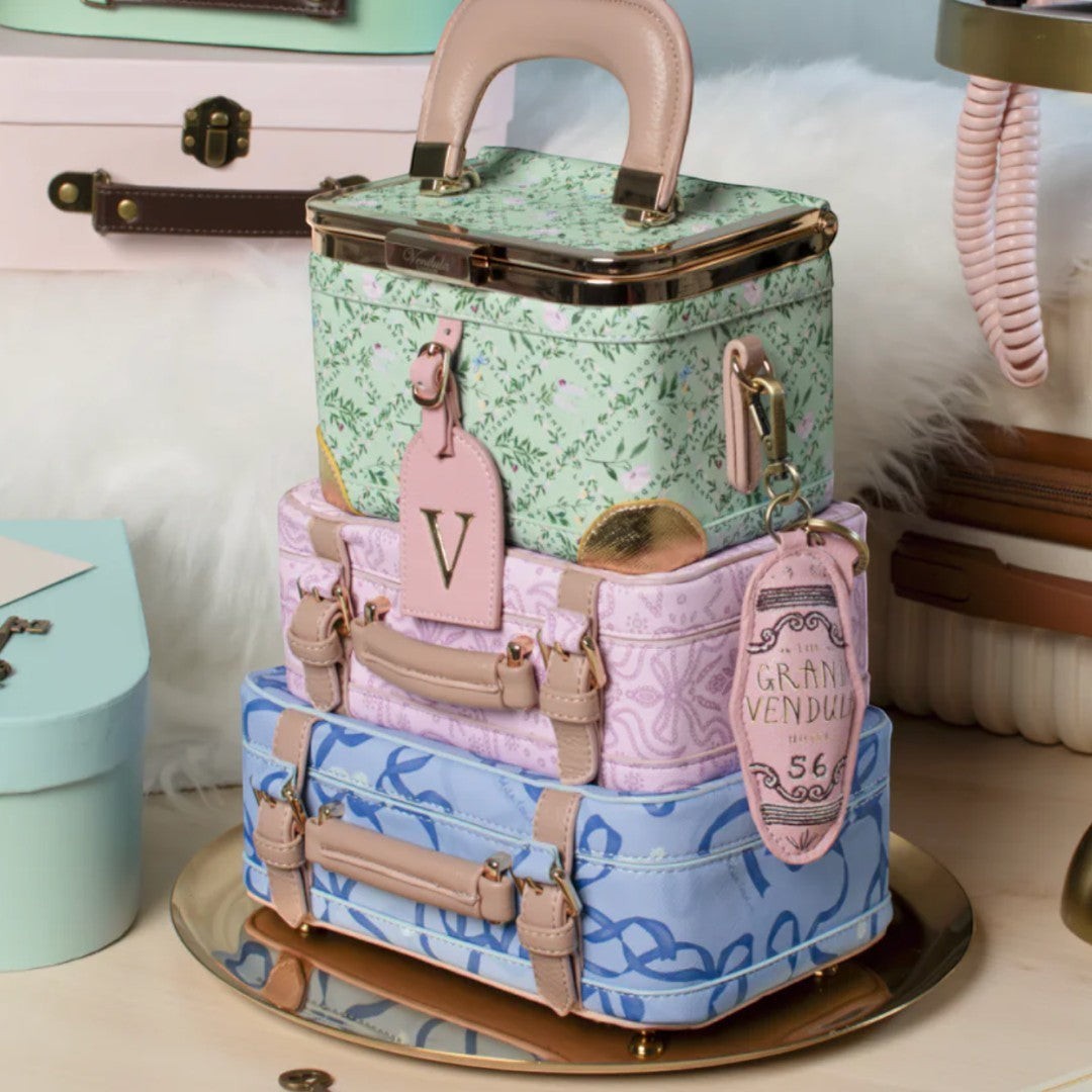 The Happy Shoe Shop Grand Vendula Hotel Key Charm, a pastel pink luggage tag with "GRAN VENDULA" and "56" printed on it, attached to a stack of miniature suitcases.