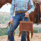 The Happy Shoe Shop Wrangler Adelyn bag featuring tooled leather, suede fringe, and logo badge, held by a woman.